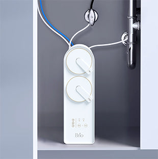 A white Brio water filtration system is installed under a sink, with wires and tubes connected to plumbing pipes. The device has two round dials and labeled buttons on its front panel.